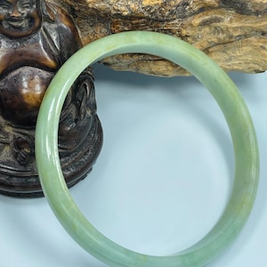 May include: A light green jade bangle bracelet. The smooth, circular bracelet is a pale green color with subtle variations in tone. The bracelet is displayed against a light blue background with a wooden carving in the background.