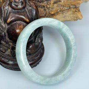 May include: A pale green jade bangle bracelet. The smooth, circular bracelet is displayed in front of a carved wooden statue of a seated Buddha. The jade has a translucent quality, with subtle variations in color.