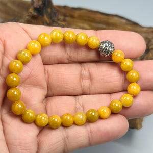 Pode incluir: Uma pulseira de contas com contas redondas amarelo dourado. A pulseira tem uma conta prateada com um padr&atilde;o como ponto focal. As contas s&atilde;o enfiadas em uma faixa el&aacute;stica. A pulseira &eacute; mostrada em uma m&atilde;o.