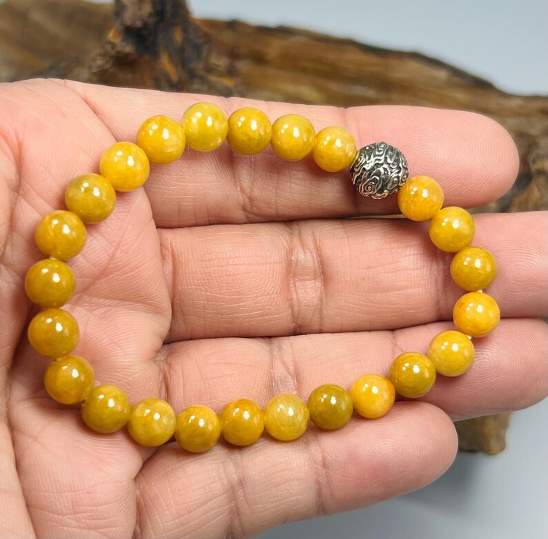 Pode incluir: Uma pulseira de contas com contas redondas amarelo dourado. A pulseira tem uma conta prateada com um padr&atilde;o como ponto focal. As contas s&atilde;o enfiadas em uma faixa el&aacute;stica. A pulseira &eacute; mostrada em uma m&atilde;o.