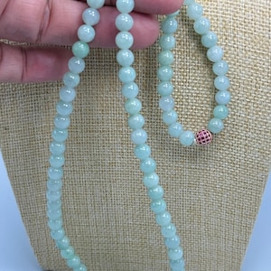 May include: A light green beaded necklace and matching bracelet. The necklace and bracelet are made of round, smooth beads. The bracelet features a small, round, red and gold accent bead. The jewelry is displayed on a neutral-colored surface.