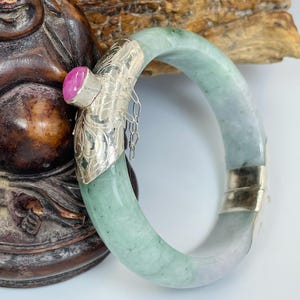 May include: A jade bangle bracelet with a silver clasp and a pink gemstone. The bracelet is a light green colour with some purple hues. The silver clasp has an ornate design and a small chain.