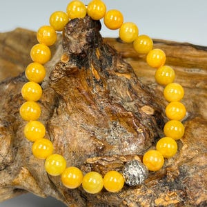 Pode incluir: Uma pulseira de contas com contas esf&eacute;ricas amarelas brilhantes. As contas s&atilde;o enfiadas juntas, com uma conta decorativa prateada como ponto focal. A pulseira &eacute; exibida em uma superf&iacute;cie de madeira marrom texturizada.