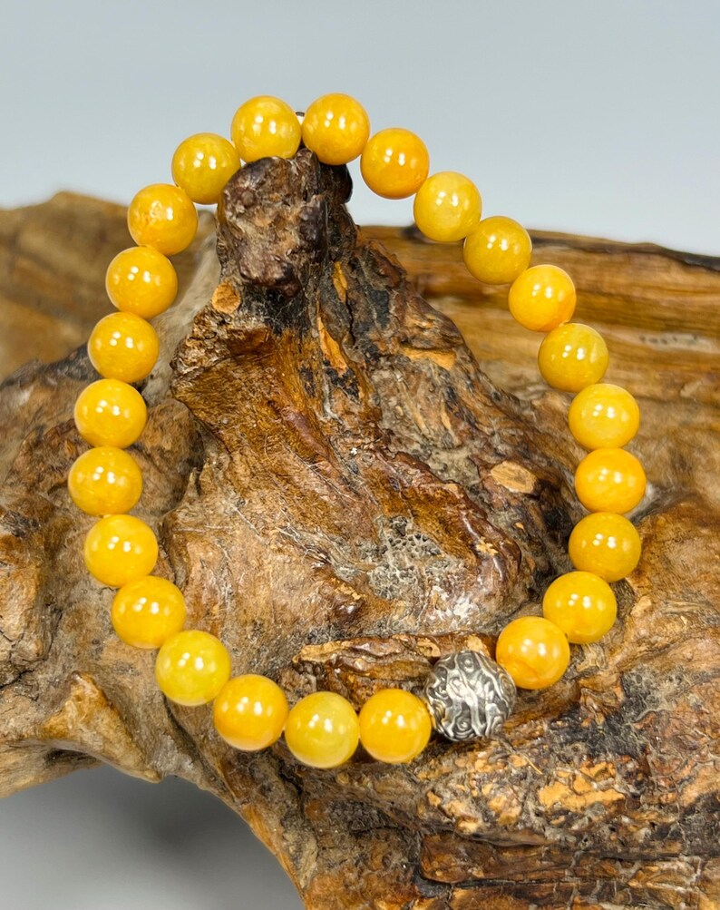 Pode incluir: Uma pulseira de contas com contas esf&eacute;ricas amarelas brilhantes. As contas s&atilde;o enfiadas juntas, com uma conta decorativa prateada como ponto focal. A pulseira &eacute; exibida em uma superf&iacute;cie de madeira marrom texturizada.