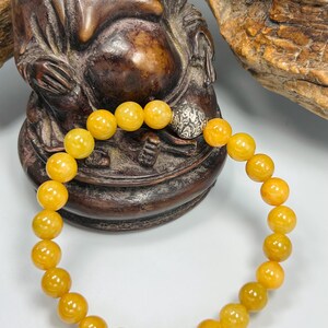 Pode incluir: Uma pulseira de contas redondas amarelo-alaranjadas. A pulseira tem uma conta prateada com um design ornamentado. &Eacute; exibida em frente a uma est&aacute;tua de madeira castanha de uma figura sentada. As contas t&ecirc;m cerca de 1 cm de di&acirc;metro.