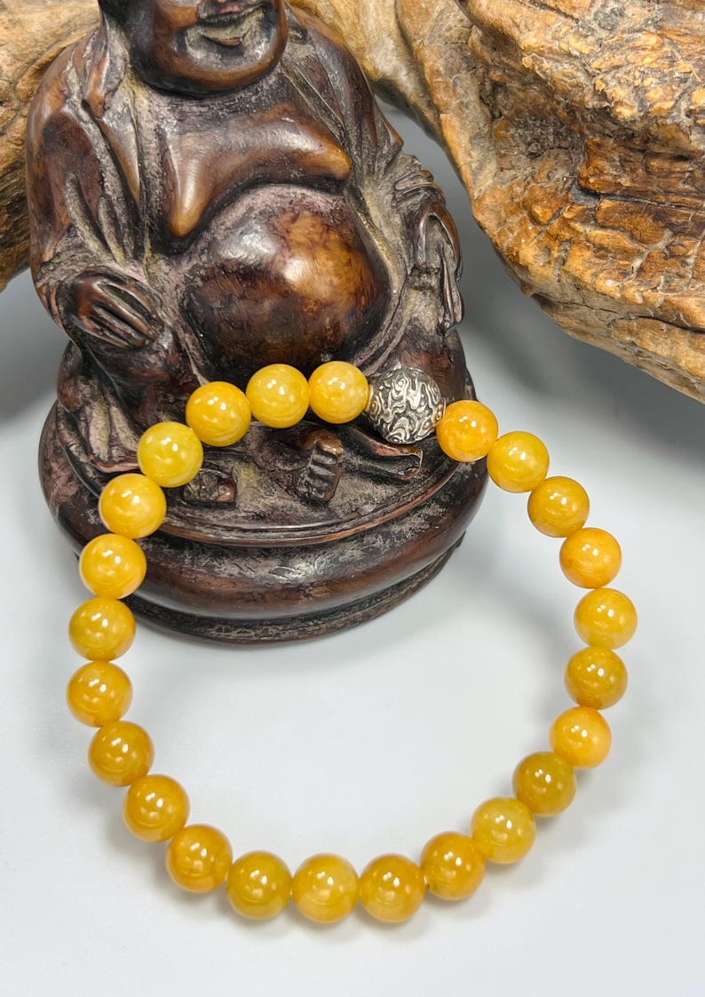 Pode incluir: Uma pulseira de contas redondas amarelo-alaranjadas. A pulseira tem uma conta prateada com um design ornamentado. &Eacute; exibida em frente a uma est&aacute;tua de madeira castanha de uma figura sentada. As contas t&ecirc;m cerca de 1 cm de di&acirc;metro.
