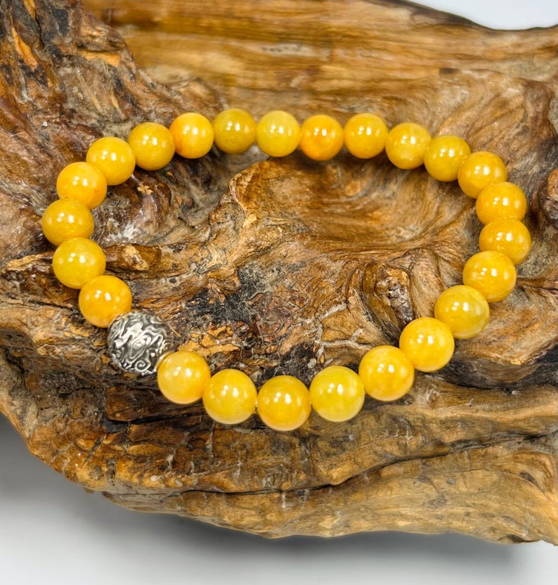 Pode incluir: Uma pulseira de contas esf&eacute;ricas amarelas brilhantes. Inclui uma &uacute;nica conta de prata ornamentada. A pulseira &eacute; exibida em uma superf&iacute;cie de madeira marrom texturizada.