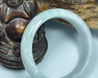 May include: A light green jade bangle bracelet. The bracelet is smooth and polished, with a translucent appearance. The jade has a subtle, natural color variation. The bracelet is displayed near a brown wooden statue of a smiling Buddha.