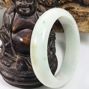 May include: A pale green jade bangle bracelet. The smooth, polished bracelet has a rounded shape and is set against a background of a bronze statue and a wooden carving.