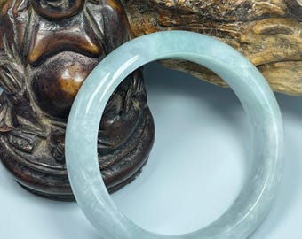 May include: A light green jadeite bangle bracelet. The smooth, polished bracelet is a pale, translucent green. The bracelet is displayed in front of a wooden carving of a smiling Buddha statue and a piece of wood.