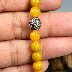 Pode incluir: Uma pulseira com contas amarelas e uma conta decorativa prateada. A pulseira &eacute; feita de contas amarelas redondas e polidas, com uma conta prateada detalhada no centro. A pulseira &eacute; segurada em uma m&atilde;o.