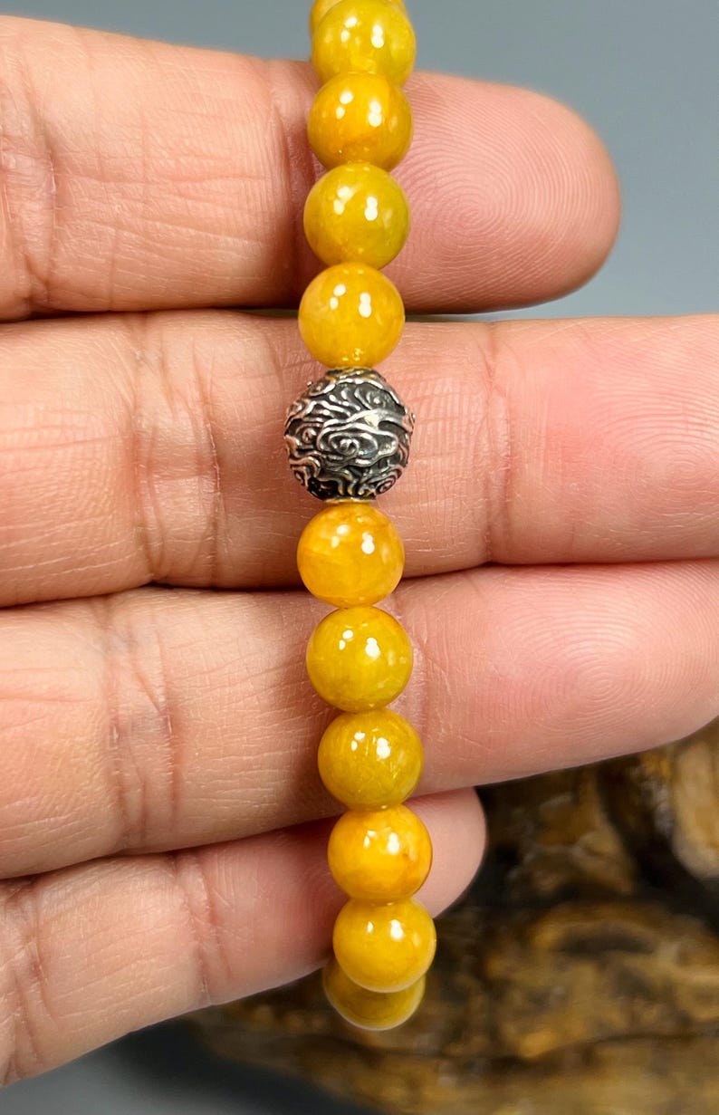 Pode incluir: Uma pulseira com contas amarelas e uma conta decorativa prateada. A pulseira &eacute; feita de contas amarelas redondas e polidas, com uma conta prateada detalhada no centro. A pulseira &eacute; segurada em uma m&atilde;o.