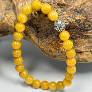 Pode incluir: Uma pulseira de contas redondas amarelas. A pulseira tem uma conta de destaque prateada com um padr&atilde;o detalhado. As contas s&atilde;o de uma cor amarelo dourado quente, e a pulseira &eacute; exibida em um fundo de madeira natural.
