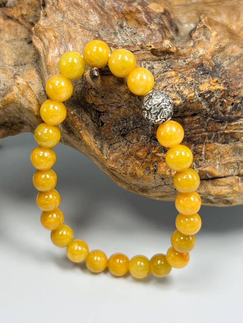 Pode incluir: Uma pulseira de contas redondas amarelas. A pulseira tem uma conta de destaque prateada com um padr&atilde;o detalhado. As contas s&atilde;o de uma cor amarelo dourado quente, e a pulseira &eacute; exibida em um fundo de madeira natural.