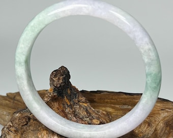 May include: A jade bangle bracelet with a smooth, polished surface. The bracelet features a gradient of colors, transitioning from a pale green to a white hue. The bangle is displayed on a piece of textured wood.