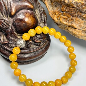 Pode incluir: Uma pulseira de contas amarelas com fecho prateado. A pulseira apresenta contas redondas e polidas em tons de amarelo e laranja. A pulseira &eacute; exibida contra um fundo de uma est&aacute;tua de madeira e uma superf&iacute;cie clara.