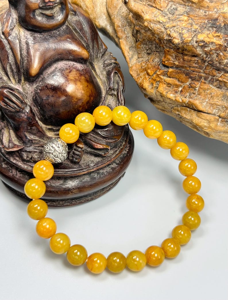 Pode incluir: Uma pulseira de contas amarelas com fecho prateado. A pulseira apresenta contas redondas e polidas em tons de amarelo e laranja. A pulseira &eacute; exibida contra um fundo de uma est&aacute;tua de madeira e uma superf&iacute;cie clara.