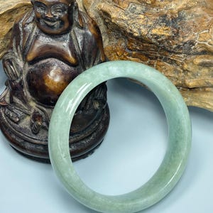 May include: A light green jade bangle bracelet. The bracelet is circular and has a smooth, polished surface. The background includes a wooden Buddha statue and a piece of wood.