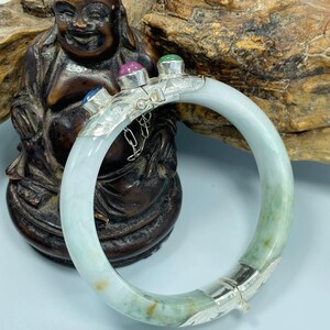 May include: A circular jade bangle bracelet featuring a silver hinge and clasp mechanism. The silver section is adorned with three small, round, cabochon-cut gemstones in blue, pink, and green. The jade band displays pale green and white tones.