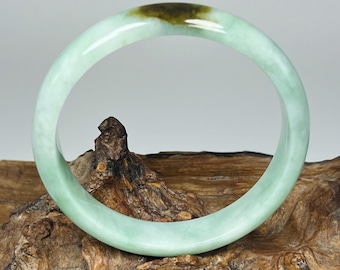 May include: A jade bangle bracelet with a light green hue and a darker green section. The smooth, circular bracelet is displayed on a piece of textured, brown wood. The bangle is a piece of jewelry.