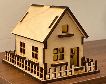 May include: A wooden model of a house with a white picket fence. The house has a brown roof, two windows on the side, and a door on the front. The house is sitting on a wooden surface.