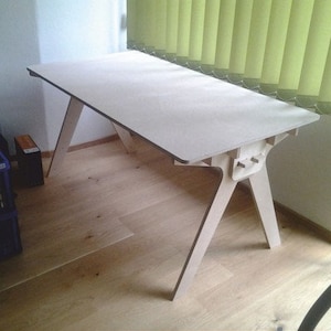 Könnte beinhalten: Ein helles Holzschreibtisch mit abgewinkelten Beinen. Der Schreibtisch hat eine rechteckige Tischplatte und ein minimalistisches Design. Die Beine sind mit einer einzigartigen Verbindung an der Tischplatte befestigt. Der Schreibtisch ist ca. 152 cm lang und 76 cm breit.