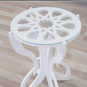 May include: White side table with a round glass top and a geometric design cut out of the wood. The table has a unique, intricate design with four legs.