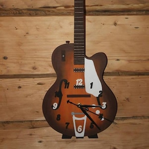 May include: A guitar-shaped wall clock in brown and orange tones. The clock face has black numerals and hands, with a white pickguard. The clock is mounted on a wooden wall.