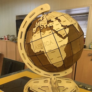 May include: A wooden globe with a stand. The globe is made of light brown wood and has a dark brown stain. The globe is labeled with continents and countries. The stand is made of light brown wood and has a dark brown stain. The globe is on a yellow surface.