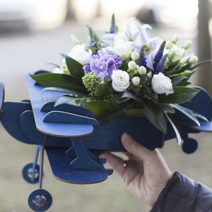 May include: A blue wooden airplane with a bouquet of white and purple flowers in the center. The airplane is a decorative piece for a flower arrangement.