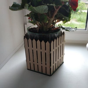 May include: A red flowering plant in a glass pot with a wooden picket fence planter. The plant is on a white windowsill.