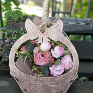 May include: A wooden basket with a bow and rope handle, decorated with a rustic brown finish. The basket is filled with pink flowers.