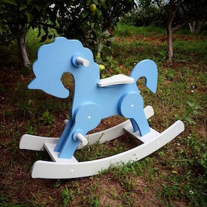 Op de afbeelding: Een lichtblauw houten schommelpaard met witte accenten. Het paard heeft een gestileerde manen en staart, een witte zitting en witte schommelpoten. Het speelgoed is ontworpen voor jonge kinderen en staat buiten in een grasveld.