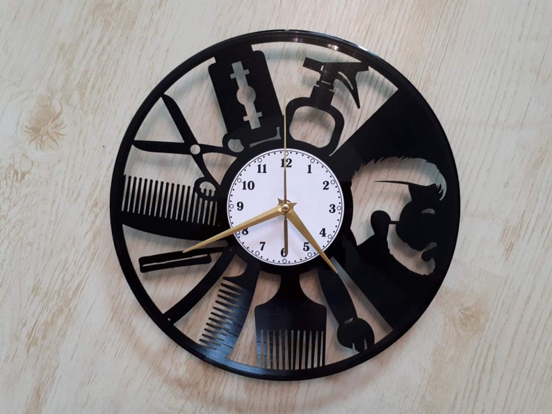 May include: Black vinyl record clock with a white clock face and gold hands. The clock is decorated with a silhouette of a barber's tools, including scissors, combs, razors, and a hairspray bottle.