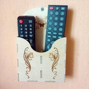 May include: A wooden wall-mounted remote control holder with two black remotes inside. The holder has a decorative floral design.