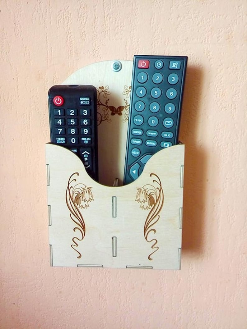 May include: A wooden wall-mounted remote control holder with two black remotes inside. The holder has a decorative floral design.