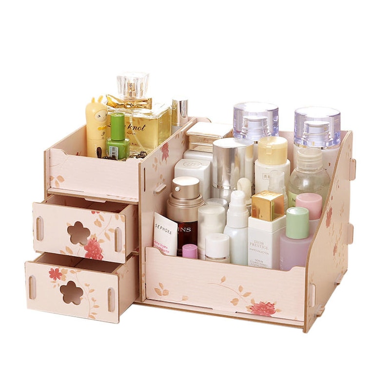 May include: A pink wooden organiser with two drawers and a top shelf for storing cosmetics and toiletries. The organiser has a floral design and is filled with various bottles and containers.