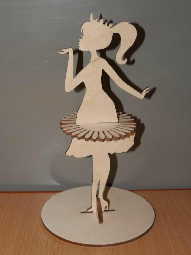 Puede incluir: Silueta de madera de una mujer con un vestido, soplando un beso. La figura tiene una corona, una cola de caballo y una falda tipo tut&uacute;. La figura de madera est&aacute; sobre una base redonda.