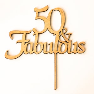 Puede incluir: Topper de pastel de madera con el texto "50 & Fabulous" en una fuente cursiva. El número 50 es más grande que la palabra "Fabulous".