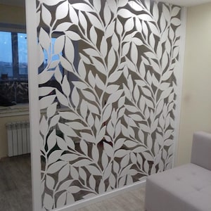 May include: White room divider with a cut-out design of leaves and branches. The divider is made of wood and is standing in a room with a white sofa.