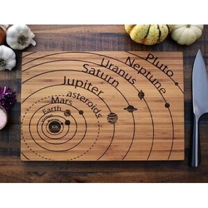 May include: A wooden cutting board with a laser-engraved design of the solar system. The board features the planets Mercury, Venus, Earth, Mars, Jupiter, Saturn, Uranus, Neptune, and Pluto, as well as the Sun and asteroid belt. The board is made of bamboo and has a natural wood finish.
