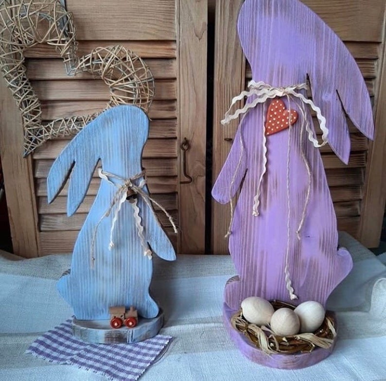 Puede incluir: Dos figuritas de conejo de Pascua de madera, una pintada de azul claro y la otra de lavanda. El conejo azul tiene un peque&ntilde;o cami&oacute;n de juguete en su base. El conejo lavanda tiene un nido de paja con tres huevos blancos.