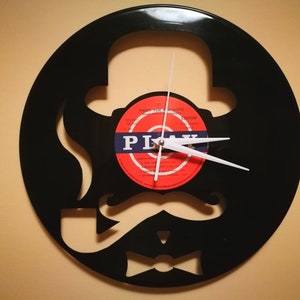 May include: A black vinyl record clock with a silhouette of a person wearing a hat, bow tie, and mustache. The clock face is a red and white record label with the text "PLAY" in blue.