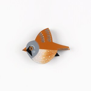 Könnte beinhalten: Ein kleiner, orangefarbener und grauer Vogel mit weißen Punkten, auf Holz gemalt. Der Vogel hat ein schwarzes Auge mit gelbem Zentrum und einen silbernen Schnabel.