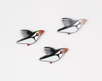 May include: Three wooden bird ornaments in the shape of puffins. The puffins are painted white with black wings and orange beaks. They are flying in different directions.
