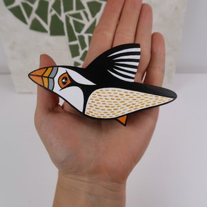 May include: A hand-painted wooden bird ornament in the shape of a puffin. The puffin is black, white, and orange with yellow accents. The bird is flying with its wings spread out.