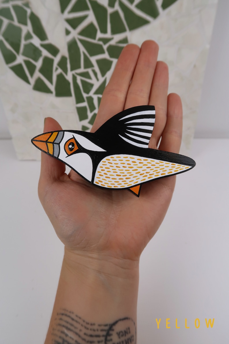 May include: A hand-painted wooden bird ornament in the shape of a puffin. The puffin is black, white, and orange with yellow accents. The bird is flying with its wings spread out.