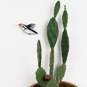 May include: A black and white wooden bird figurine with orange and red accents, flying in front of a green cactus plant in a brown pot.