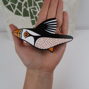 May include: A hand holds a wooden bird carving of a puffin. The puffin is painted black, white, orange, and brown. The bird is flying with its wings spread out.