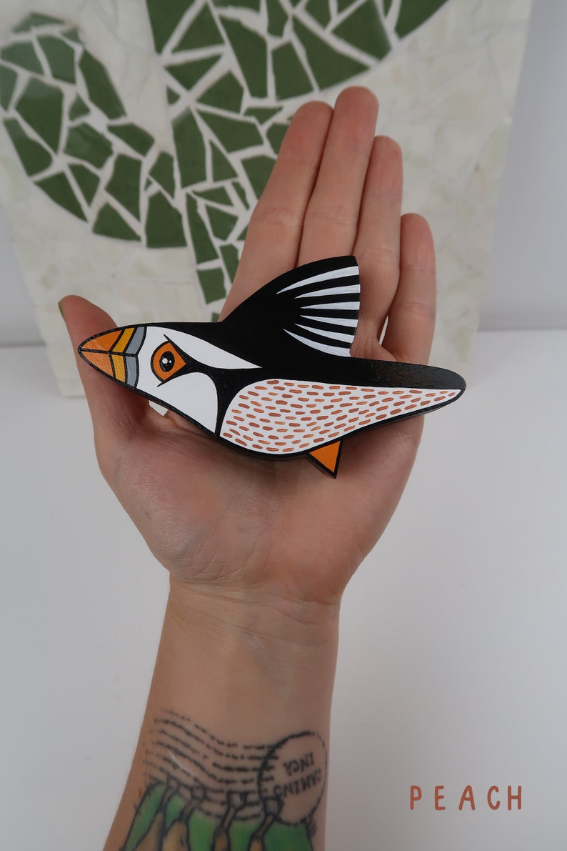 May include: A hand holds a wooden bird carving of a puffin. The puffin is painted black, white, orange, and brown. The bird is flying with its wings spread out.